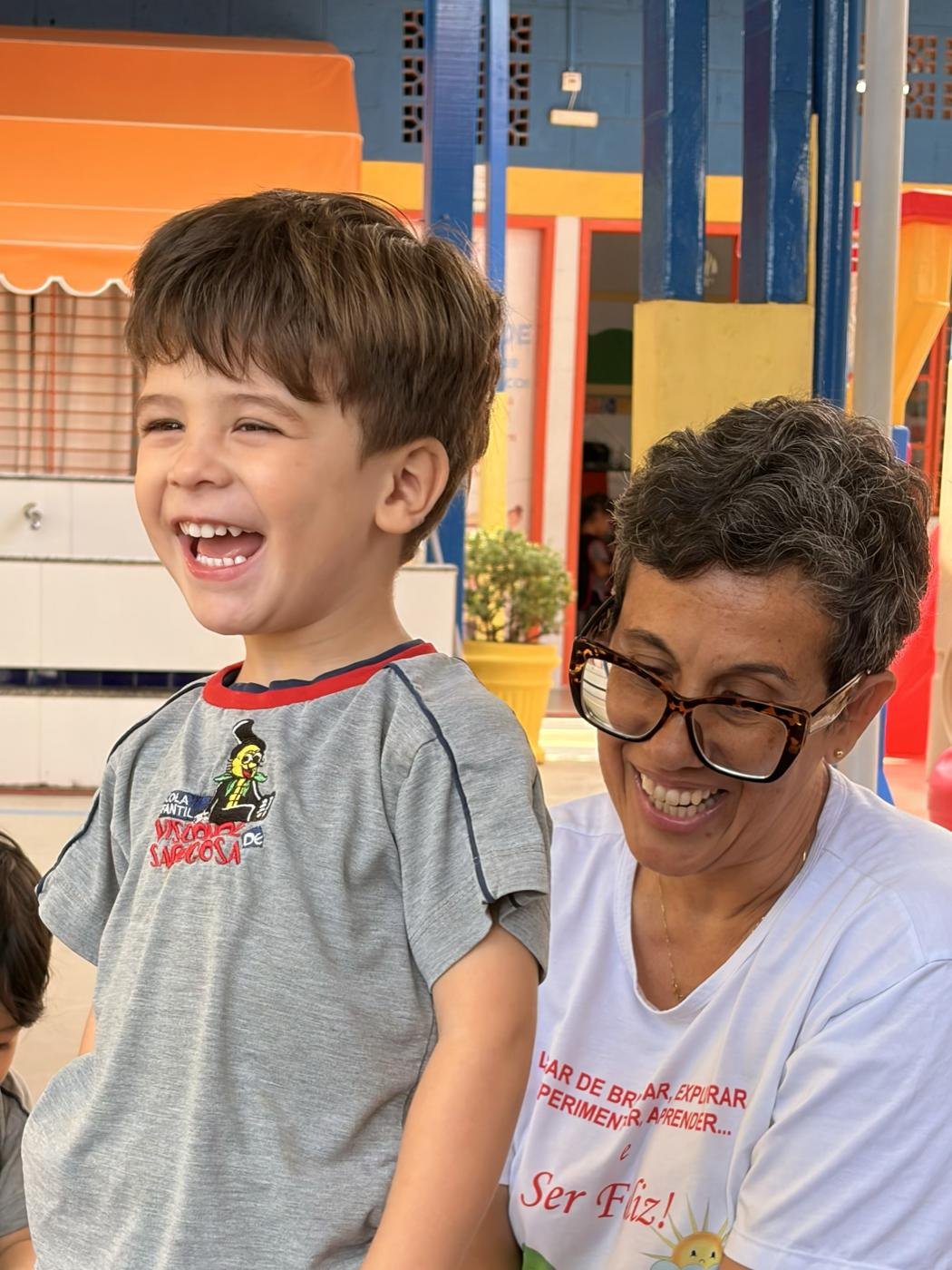 Menino rindo ao lado de uma professora no pátio da Escola Visconde de Sabugosa
